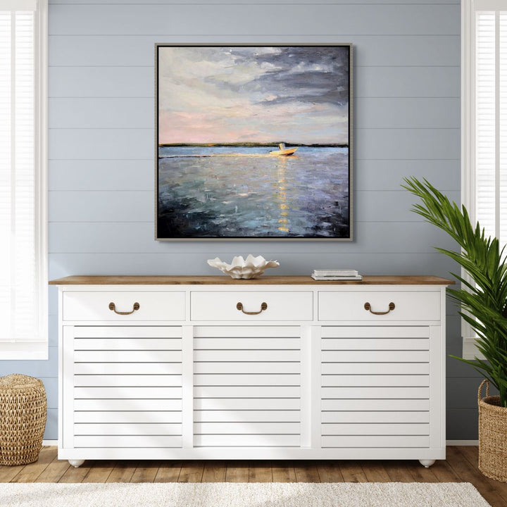 White cabinet with wooden top against a gray wall with a framed painting of a sunset over water.